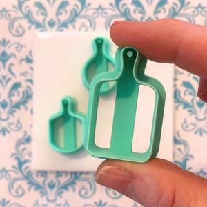 May include: A set of three teal-colored, miniature cutting board-shaped cookie cutters. One cutter is held by a person's fingers, with two others visible in the background. The cutters have a rectangular shape with a handle and a white stripe.
