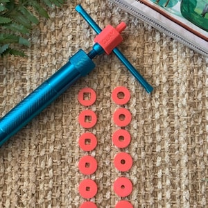May include: A blue clay extruder tool with a red handle and the text "CLAY BOX". The tool is accompanied by several coral-colored clay shape cutters, including squares and circles. The items are arranged on a textured, beige surface.