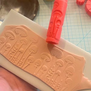 May include: A red mushroom-themed clay roller and a clay stamp with a mushroom design. The stamp is pressed into a piece of light orange clay, creating an embossed pattern. Other clay tools and a jar of gold powder are in the background.