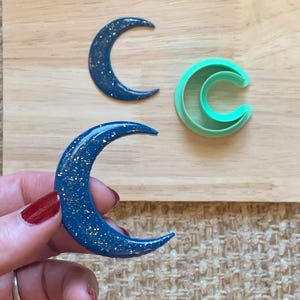 May include: Three crescent moon-shaped items. Two are dark blue with silver flecks, one is held by a hand with red nail polish. The other is a green plastic cookie cutter. All are on a wooden surface.