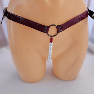 May include: A burgundy leather belt with a silver metal ring and a strand of white pearls. The belt has silver metal hardware and is displayed on a mannequin. The pearls hang down from the ring.