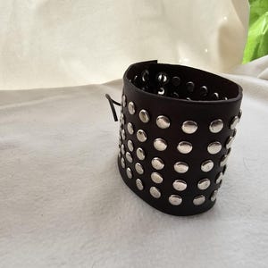 Może przedstawiać: Bransoletka ze skóry w kolorze czarnym z srebrnymi ćwiekami. Bransoletka ma około 5 cm szerokości i prosty design.