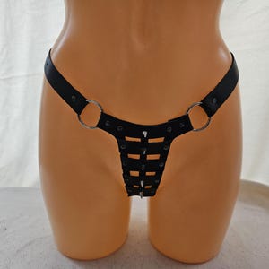 Op de afbeelding: Zwarte lederen lingerie met zilveren metalen ringen en studs. De lingerie heeft een kruislings ontwerp aan de voorkant.