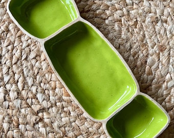 May include: A ceramic serving dish with three connected compartments. The interior of each compartment is a bright, glossy green. The dish has a rectangular shape with rounded corners and sits on a woven, tan surface.