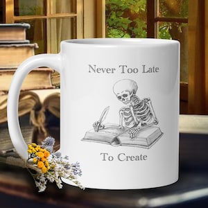 Puede incluir: Taza de café de cerámica blanca con una ilustración en blanco y negro de un esqueleto escribiendo en un libro. La taza tiene el texto "Never Too Late To Create" impreso en ella.
