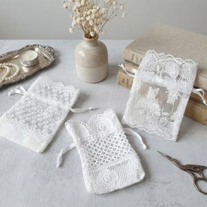 Puede incluir: Tres bolsas de encaje blanco con cierres de cordón se exhiben sobre una superficie clara. También hay una bandeja plateada con un collar de perlas y una vela, un jarrón con flores secas y libros antiguos. Unas tijeras pequeñas descansan cerca.