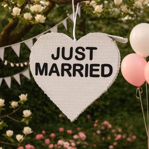 May include: A white heart-shaped piñata with the words "JUST MARRIED" in black lettering hangs from a tree branch. White and pink balloons are visible in the background, along with a garland of white flags and flowers, suggesting a wedding or celebration.