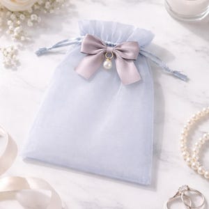 Puede incluir: Una bolsa de regalo de organza azul claro con un lazo de satén gris y un colgante de perla. La bolsa está atada con un cordón azul a juego. El fondo incluye una vela encendida, joyas y flores.