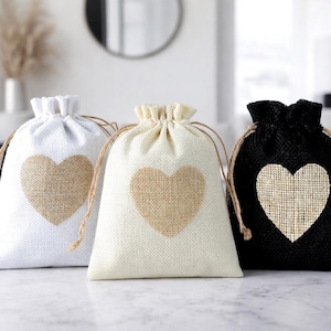 Puede incluir: Tres bolsas de regalo con cordón en blanco, crema y negro, cada una con un corazón de yute. Las bolsas tienen un cierre de cordón de yute natural. Ideales para pequeños regalos.