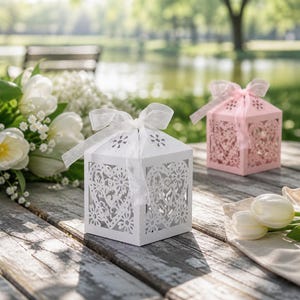 Puede incluir: Dos cajas decorativas para recuerdos, una blanca y otra rosa, sobre una superficie de madera envejecida. Las cajas tienen intrincados recortes de corazón y flores y están atadas con cintas transparentes. Flores blancas y vegetación dispuestas cerca.