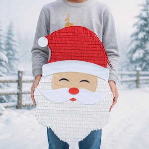 May include: A large Santa Claus piñata with a red hat, white beard, and smiling face is held in front of a person. The piñata is made of paper and has a textured appearance. The background shows a snowy winter scene.