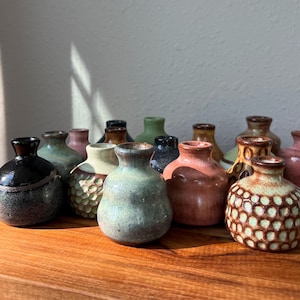 May include: A collection of small, round ceramic bud vases in various colors and textures, including glossy black, speckled green, matte pink, and honey-brown with honeycomb patterns, arranged together on a wooden surface.