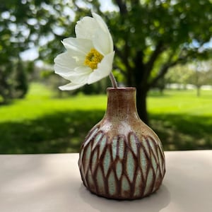 May include: A small, handcrafted ceramic vase with a brown and turquoise diamond pattern. A single white flower with a yellow center is placed inside the vase. The vase is on a light-colored surface with a blurred green background.