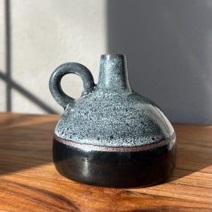 May include: A ceramic vase with a rounded body and a small handle. The top half is speckled gray and white, while the bottom half is black. A thin brown band separates the two colors. The vase is on a wooden surface.