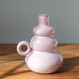May include: A pink ceramic vase with a unique, stacked design. The vase has a small handle and a speckled texture, adding visual interest. The color is a soft, dusty rose, suitable for displaying flowers or as a decorative piece.
