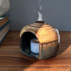 May include: A round ceramic oil burner with a glossy glaze featuring a gradient of warm yellow and deep brown tones. The piece has a wide arched opening at the front containing a small white tea light candle with a visible flame and rising smoke.