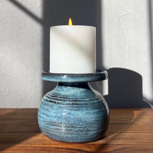 May include: A blue ceramic candle holder with a white candle. The candle holder has a round base and a flared top. The candle is lit with a yellow flame. The candle holder is on a wooden surface.