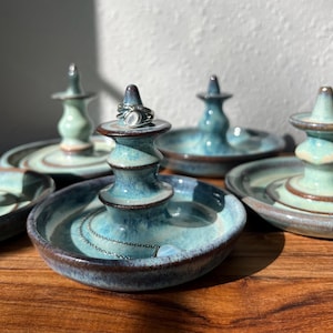 May include: A set of ceramic ring holders with a glossy blue and green glaze and dark brown rims. Each holder features a central conical post for rings and a shallow dish base. One holder displays a silver ring with a white stone and a silver chain.