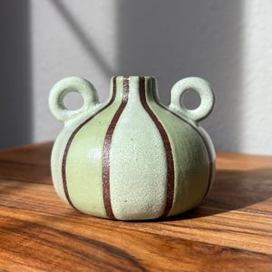May include: A small, round, light green ceramic vase with two loop handles. The vase features vertical brown stripes and a textured surface. The vase is sitting on a wooden surface.