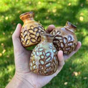 May include: Three small ceramic vases with narrow necks and rounded bodies. Each vase features a textured pattern of raised, leaf-like indentations. Two vases are glazed in shades of brown and tan, while one is glazed in light green and brown.