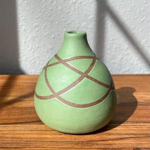 May include: A small, green ceramic vase with a rounded body and a narrow neck. The vase is decorated with a crisscross pattern in a darker, copper-colored glaze. The vase sits on a wooden surface.