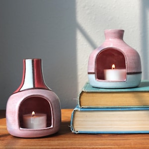 May include: Two ceramic candle holders with arched openings, each containing a lit white tea light candle. One holder features a pink and white vertical striped pattern, while the other is a solid pink and light blue color block design.