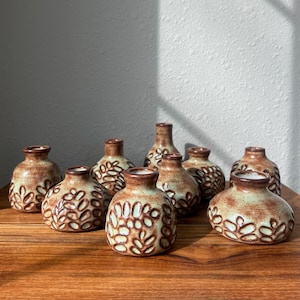 May include: A collection of small, brown ceramic vases with a leaf pattern. The vases have a light green interior and are arranged on a wooden surface. The vases vary in shape and size.