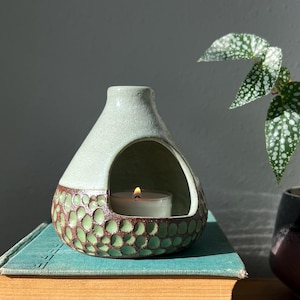 May include: A ceramic oil burner with a light green matte top and a textured, dark brown base featuring circular indentations. The burner has a wide, arched opening containing a small, lit white tea light candle, creating a warm glow.