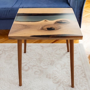 May include: Square wooden coffee table with a natural wood grain finish and a dark blue resin inlay. The table has four slender, brown legs and is set on a patterned rug. The table measures approximately 24 inches long and wide, and 18 inches high.