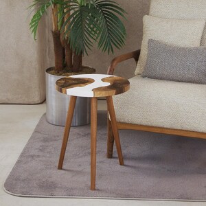 May include: A small, round side table with a wooden top and three wooden legs. The tabletop features a natural wood grain pattern with a white resin inlay. The table is placed on a gray rug near a cream-colored armchair.