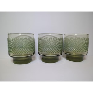 May include: Three vintage green-tinted drinking glasses. Each glass has a thick, rounded base and a cylindrical body with a wavy, etched design. The glasses are made of glass and are suitable for serving beverages.