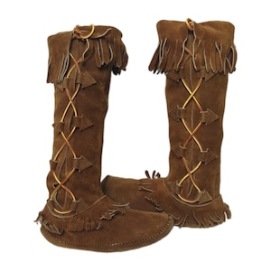 May include: A pair of knee-high brown suede boots featuring fringe detailing at the top and base. The front of each boot is decorated with a crisscross lacing pattern held by triangular suede tabs, creating a distinct textured appearance.