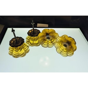 Könnte beinhalten: Vier gelbe, blütenförmige Schrankknöpfe aus Glas mit bronzefarbener Metallbeschlägen. Die Knöpfe haben ein geripptes Design und sind auf einer weißen Oberfläche angeordnet. Die Hardware umfasst eine Gewindestange und eine dekorative Basis.