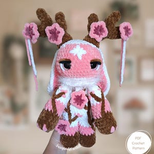 Könnte beinhalten: Handgemachte Häkelpuppe in Rosa und Weiß mit braunen Geweihen und rosa Blumenakzenten. Die Puppe hat große schwarze Augen und wird von einer Hand gehalten. Es ist ein PDF-Häkelmuster.