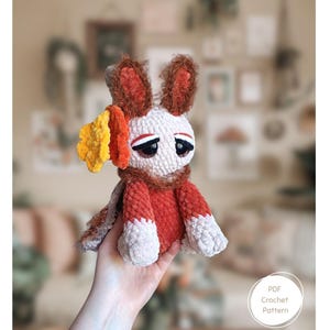 Puede incluir: Una criatura de ganchillo de felpa con cara blanca, cuerpo color óxido y orejas largas. Presenta grandes ojos oscuros con párpados pesados, un cuello marrón texturizado y un accesorio en forma de hoja amarilla y naranja. Una etiqueta circular dice PDF Crochet Pattern.