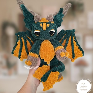 May include: A plush dragon toy crafted from dark green, mustard yellow, and light brown yarn. The creature features large wings with yellow inner panels, two horns, and a textured body. The text SassyWolfCrochet and PDF Crochet Pattern are visible.