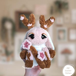 Peut inclure: Une peluche lapin crochetée avec des bois marron et des fleurs roses. Le lapin a de grands yeux noirs et tient une branche avec des fleurs roses. Le lapin est rose clair et marron. Le texte "PDF Crochet Pattern" est dans un cercle blanc.