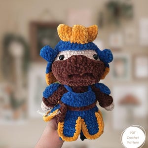 May include: A handmade crochet doll in the shape of a samurai warrior. The doll features a blue and gold helmet, armor, and accents. The face is brown with white and black details. The doll is soft and plush, with a textured appearance. The doll is a PDF crochet pattern.