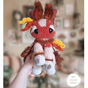 Puede incluir: Un juguete de peluche con forma de criatura hecho de hilo color naranja óxido, crema y amarillo con grandes ojos negros, orejas altas y detalles de pelaje texturizado. El juguete tiene una cabeza redondeada, extremidades pequeñas y un gráfico.