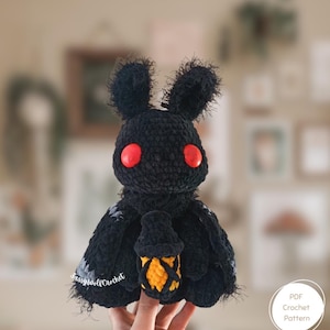 May include: A handmade black crochet moth plushie with red eyes and a small lantern. The moth has a fuzzy texture and is holding a yellow and black lantern. The text "SassyWolfCrochet" is visible on the wing.
