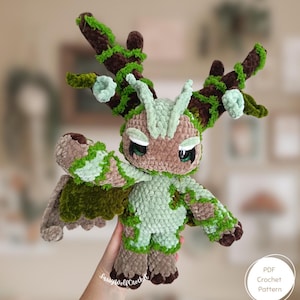 May include: A handmade crochet plush toy in the shape of a forest creature. The toy features brown, green, and light green colors, with large eyes and detailed antlers. The plush is held up by a hand. The image also includes a logo that says "PDF Crochet Pattern".