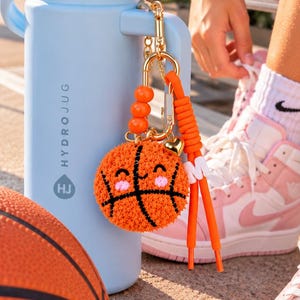 May include: A light blue HydroJug water bottle with a straw, featuring a gold-toned keychain with an orange fuzzy basketball charm, three orange beads, a small gold heart, and a white letter M charm attached to a bright orange cord.