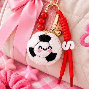 May include: A light pink quilted duffel bag featuring the embroidered name Eve and a pink bow. Attached to the bag is a gold-toned keychain with red beads, a white letter S, and a plush soccer ball charm with a smiling face and pink cheeks.