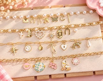 May include: Four gold-tone chain bracelets featuring various charms including pearls, a butterfly, a moon, a cowboy boot, a heart, a star, a leopard, a smiley face, a disco ball, a roller skate, and a pink flower. The bracelets are displayed on a beige board.