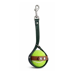 May include: A dog toy featuring a bright yellow tennis ball held in a dark green leather holder with a brown leather strap. The holder is attached to a silver metal clasp and a dark green leather strap. The brand name "Pupsense" is printed on the strap.