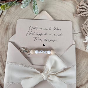 May include: A beige envelope with a cream-colored ribbon and a keychain that says "PAPI." The card inside reads "Cette année, le Père Noël t'apporte un secret... Tu vas être papi."