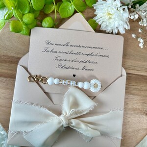 Pu&ograve; includere: Un biglietto di auguri in una busta rosa chiaro con un fiocco color crema. Il biglietto recita "Une nouvelle aventure commence... F&eacute;licitations Mamie." Un portachiavi di perline bianche con "MAMIE" &egrave; attaccato.