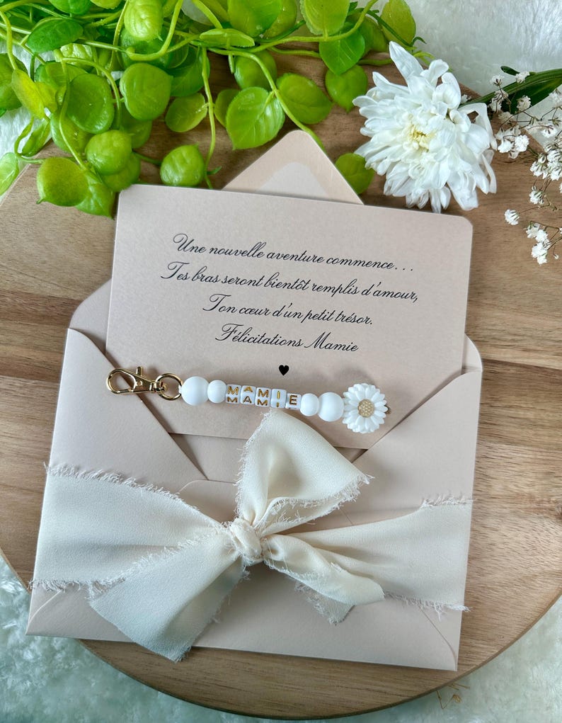 Pu&ograve; includere: Un biglietto di auguri in una busta rosa chiaro con un fiocco color crema. Il biglietto recita "Une nouvelle aventure commence... F&eacute;licitations Mamie." Un portachiavi di perline bianche con "MAMIE" &egrave; attaccato.