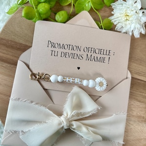 Pu&ograve; includere: Una busta beige chiaro con un nastro color crema e una carta con la scritta "PROMOTION OFFICIELLE : TU DEVIENS MAMIE !". &Egrave; incluso un portachiavi di perline bianche con la parola "MAMIE" e un ciondolo a margherita.