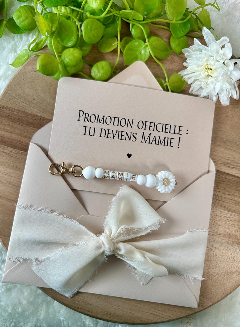 Pu&ograve; includere: Una busta beige chiaro con un nastro color crema e una carta con la scritta "PROMOTION OFFICIELLE : TU DEVIENS MAMIE !". &Egrave; incluso un portachiavi di perline bianche con la parola "MAMIE" e un ciondolo a margherita.